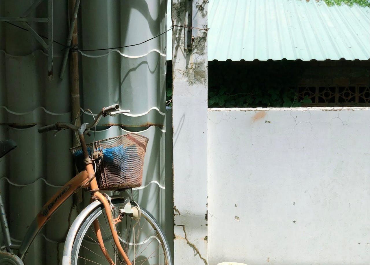 A vintage bicycle with a basket stands against a textured urban wall under sunlight.